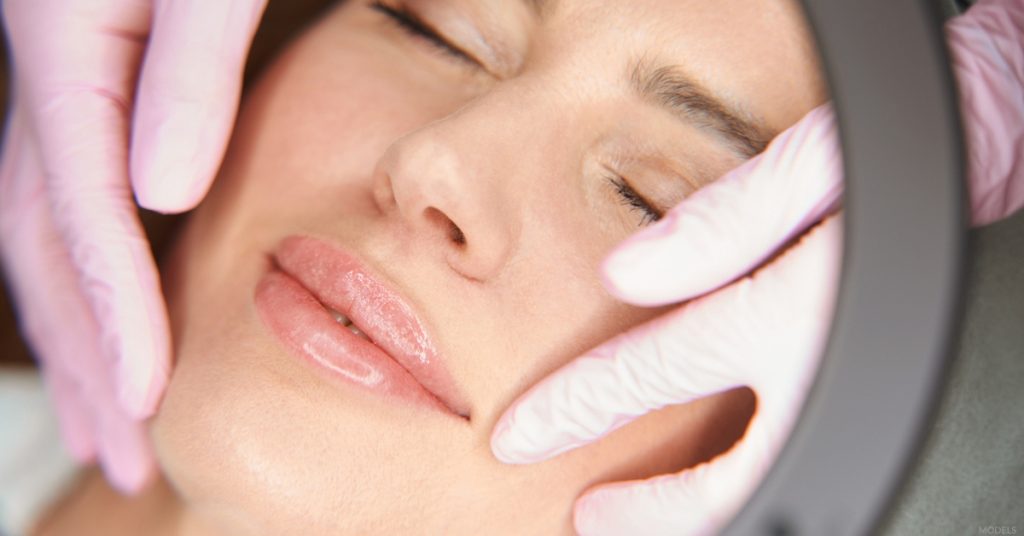 Close-up of a woman lying down with her eyes closed as an esthetician wearing pink gloves gently examines her face during a skincare treatment. (models)